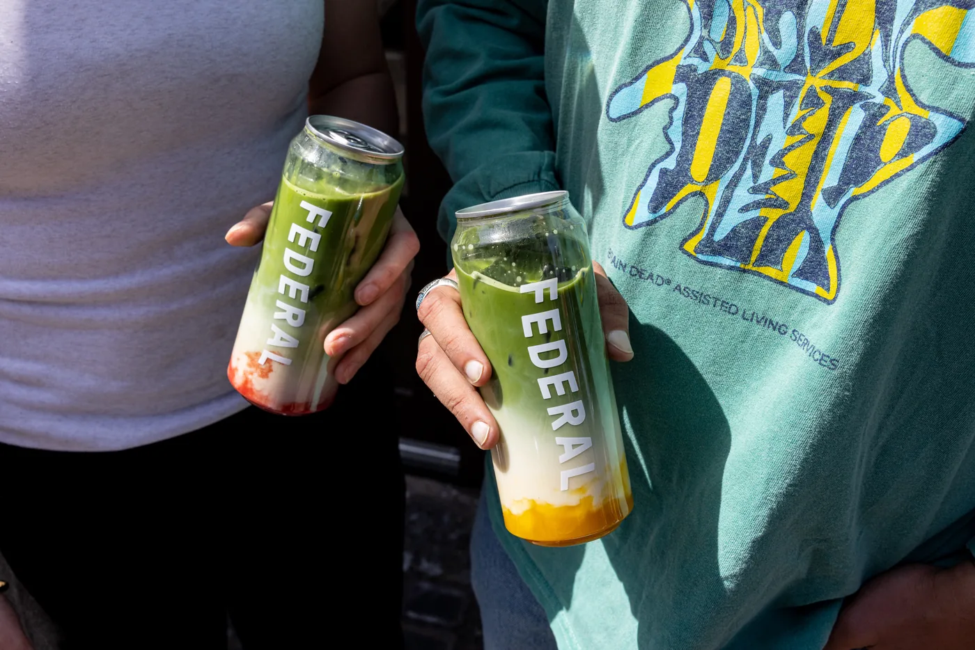 Two hands holding tall cans branded FEDERAL, each filled with layers of green matcha over milk, one with a red fruit layer at the bottom and one with a yellow layer.