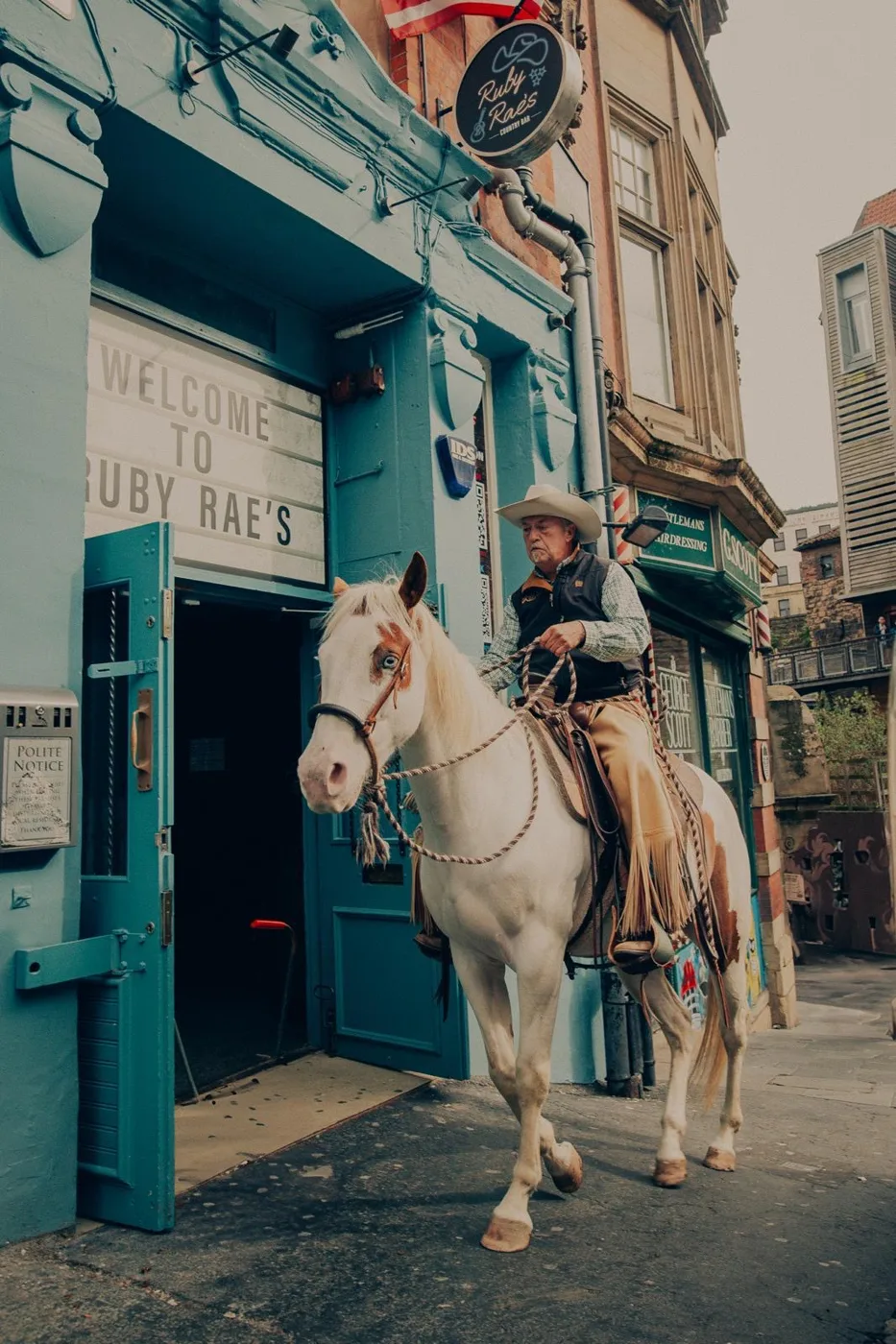 A rider in a cowboy hat on a white horse outside a teal shopfront with a sign reading Welcome to Ruby Rae's